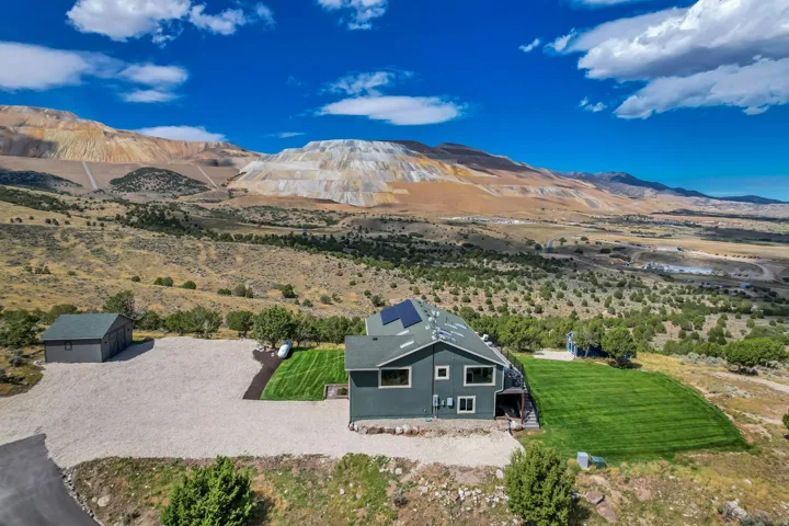 Modern residential structure with solar panels, a detached garage, expansive gravel driveway, and a manicured lawn area, set against a mountainous backdrop