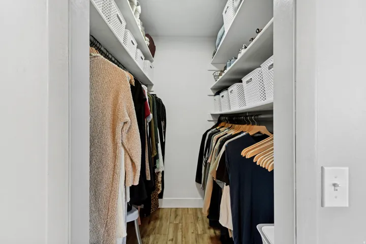 Spacious walk-in closet featuring dual clothing rods with wooden hangers, wall-mounted shelving for organized storage, wood-finish flooring, and abundant natural lighting from overhead fixtures