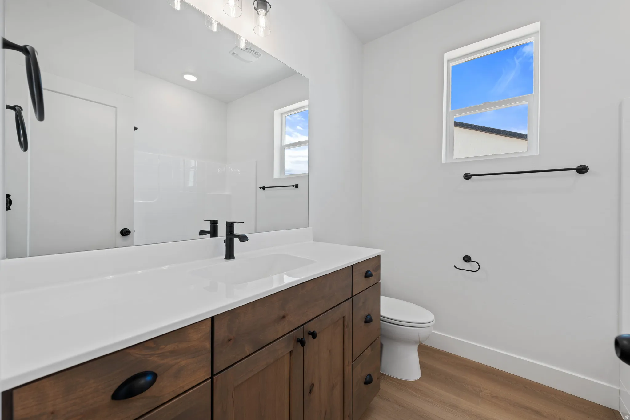 Wood-finish flooring, a wood vanity with dark hardware, an integrated sink, a large mirror, and a window