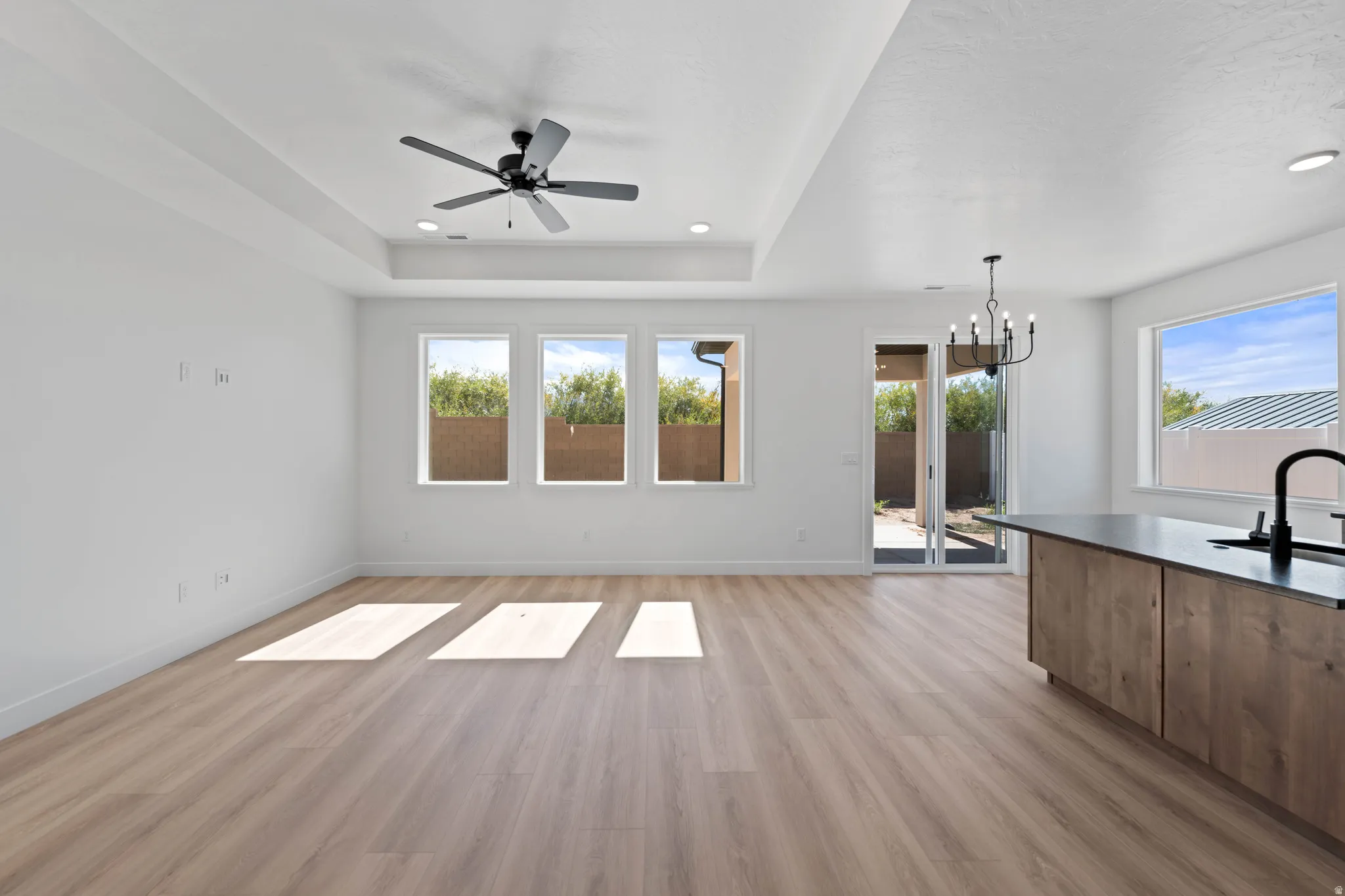 Tray ceiling with a ceiling fan, wood-finish flooring, multiple windows, kitchen island with a dark countertop and sink, and a sliding glass door to an outdoor area