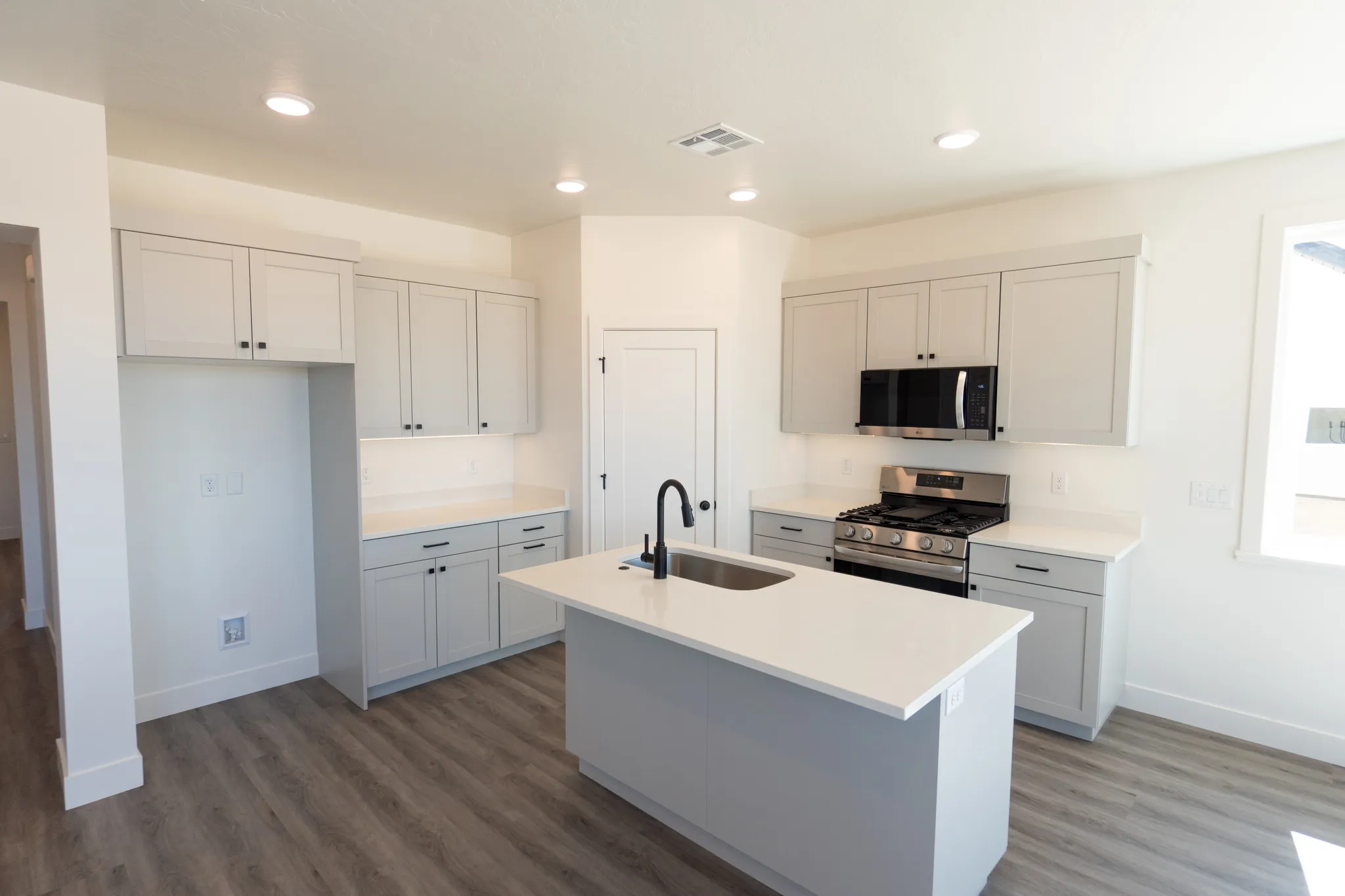 Kitchen featuring a central island with an integrated sink, light grey cabinetry, white countertops, stainless steel appliances, and wood-finish flooring