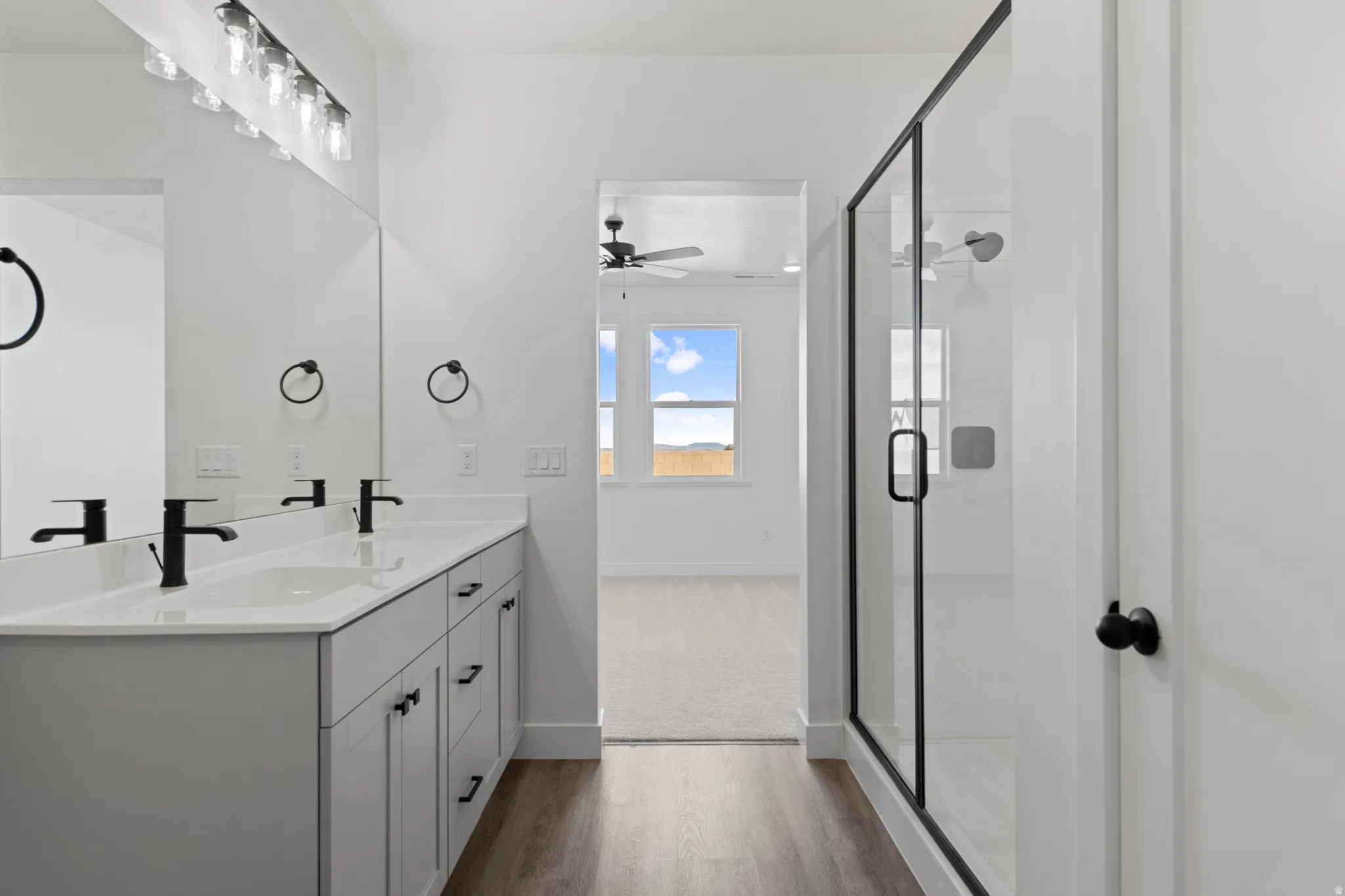 Dual vanity with matte black fixtures, framed glass shower enclosure, wood-finish flooring, and ample natural light