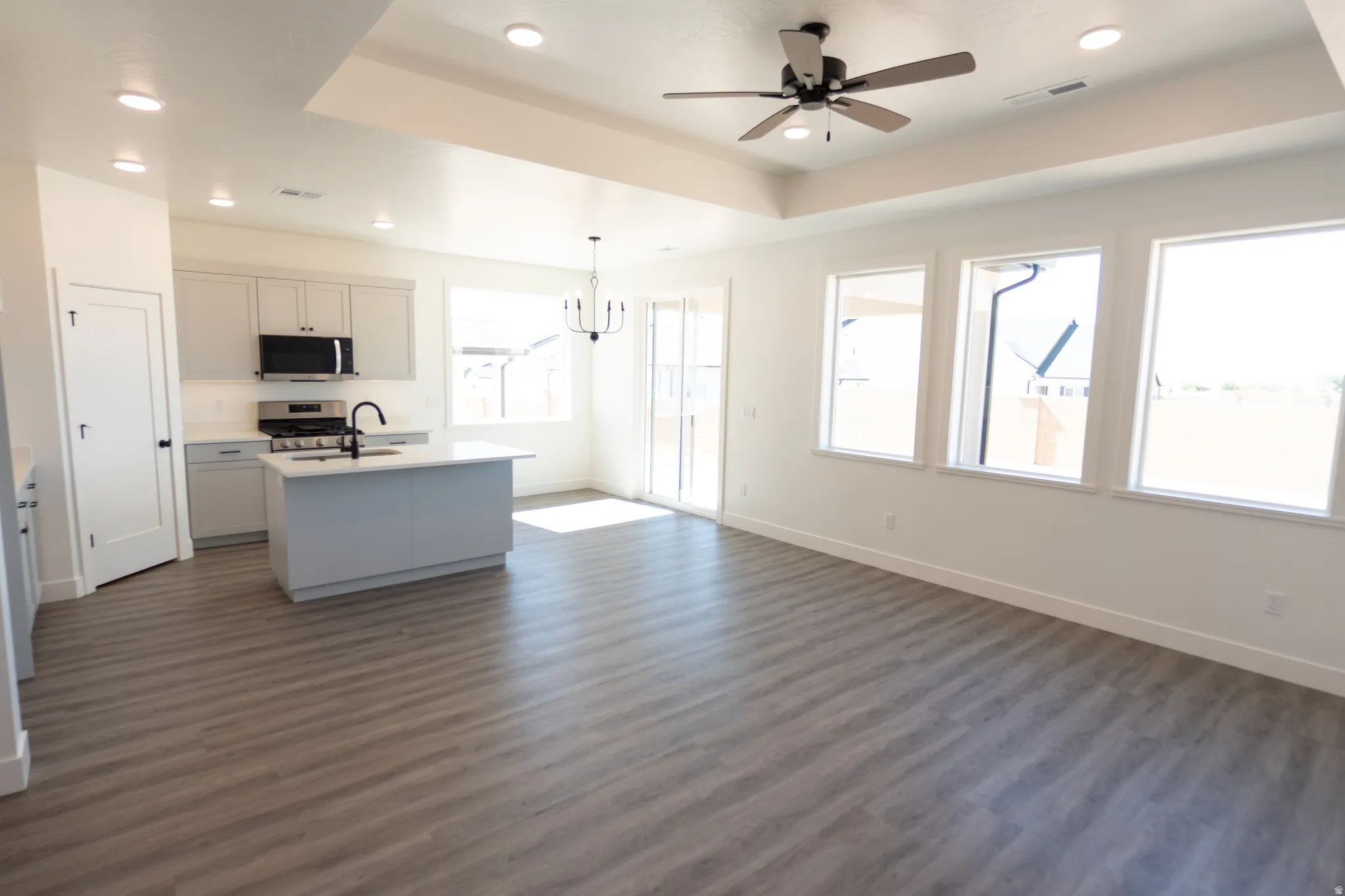 Open-concept living space with wood-finish flooring, a contemporary kitchen island, three large windows, a tray ceiling, and a ceiling fan