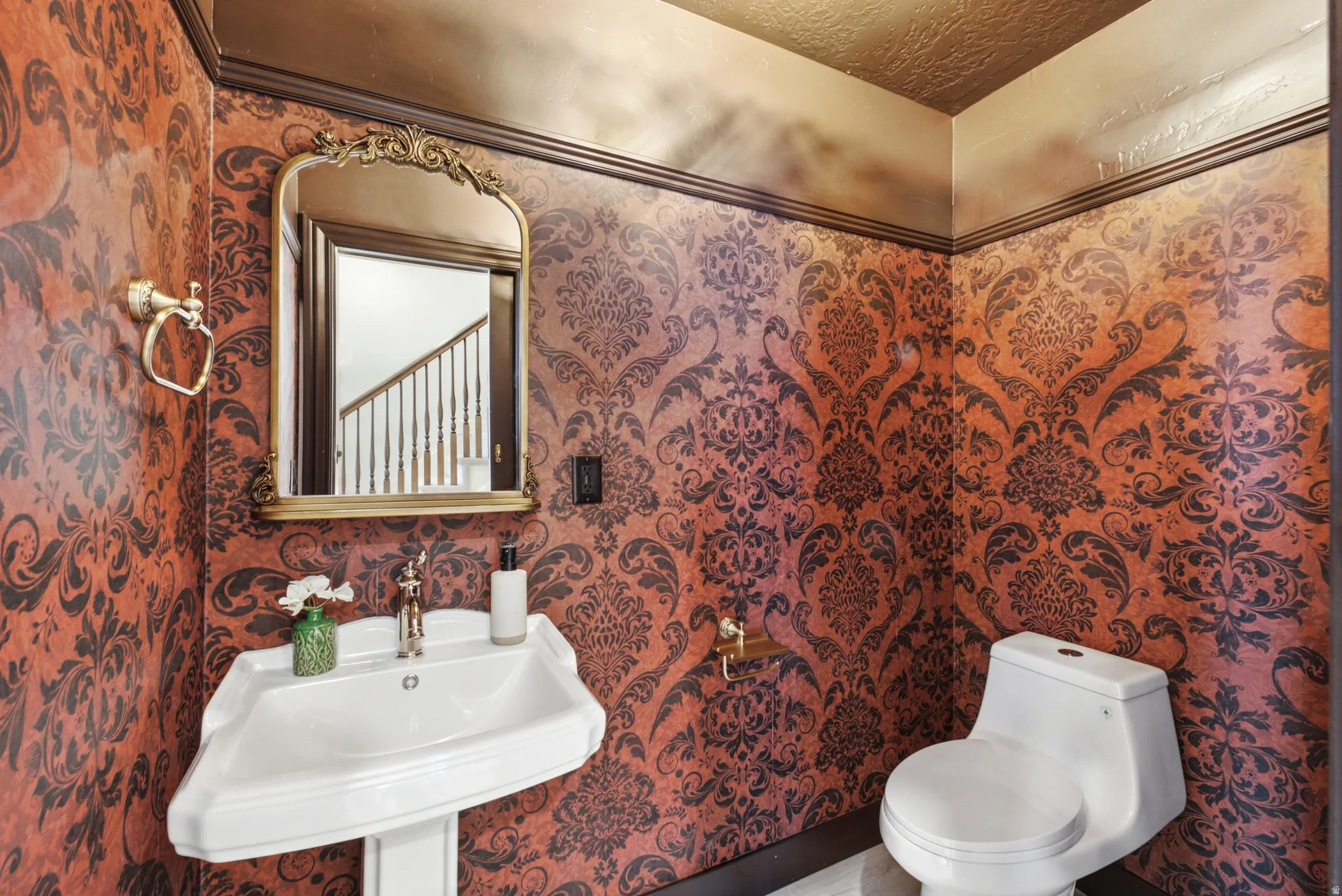 Pedestal sink with polished brass faucet, decorative framed mirror, and ornate patterned wallpaper