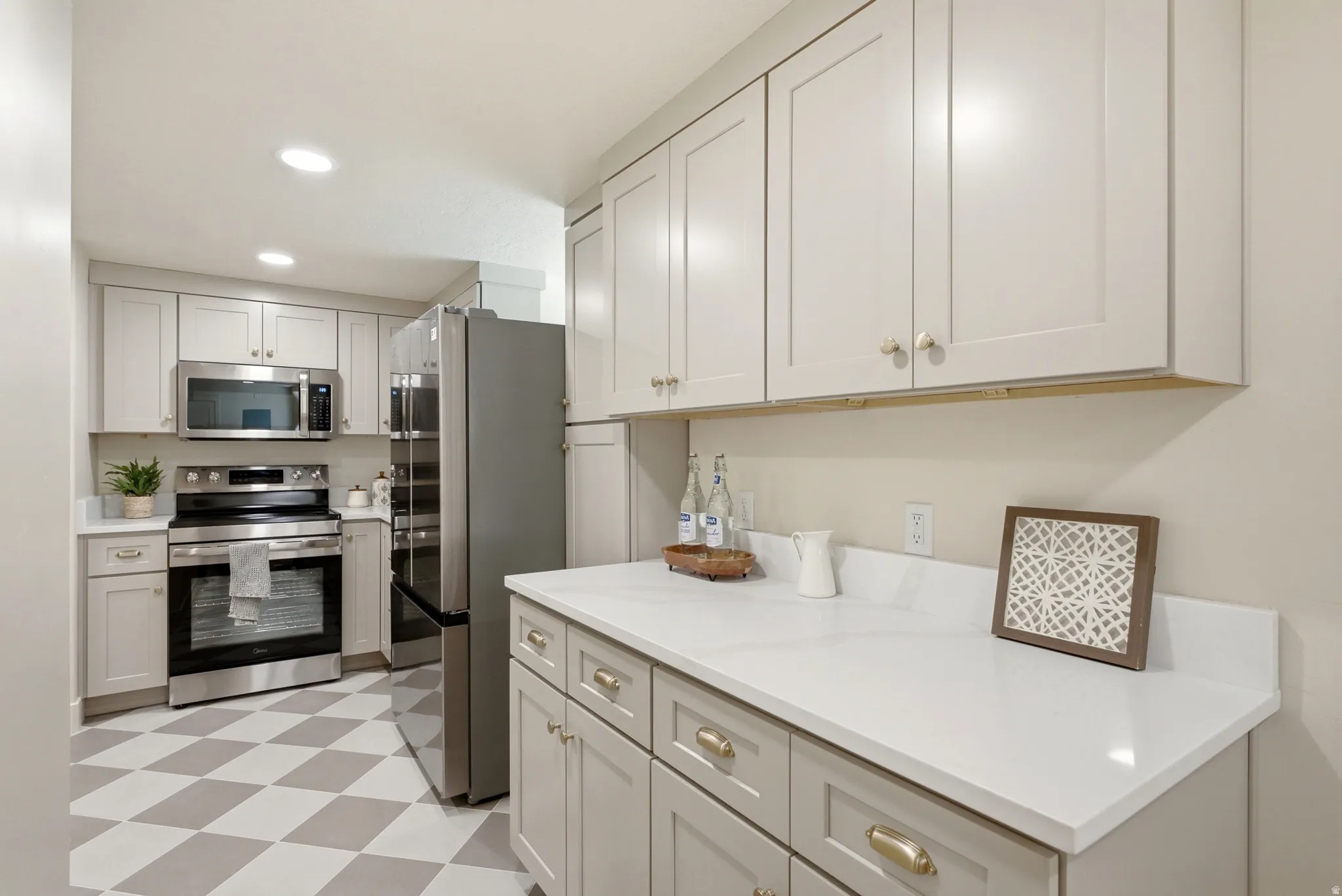 Recessed lighting illuminating light-toned cabinetry, stainless steel appliances, white countertops, and a distinct checkered floor pattern