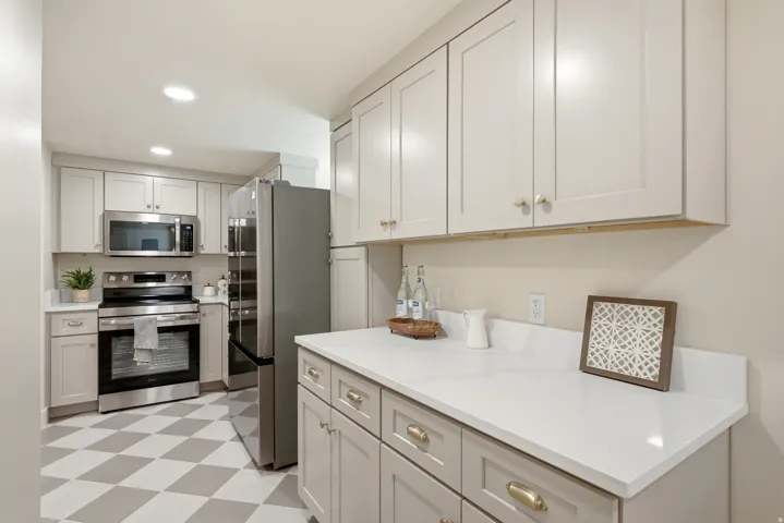Recessed lighting illuminating light-toned cabinetry, stainless steel appliances, white countertops, and a distinct checkered floor pattern