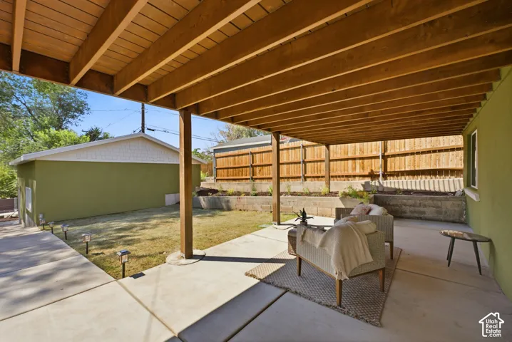 Covered concrete patio with exposed wood ceiling beams, adjacent to a grassy yard with perimeter fencing and tiered planters