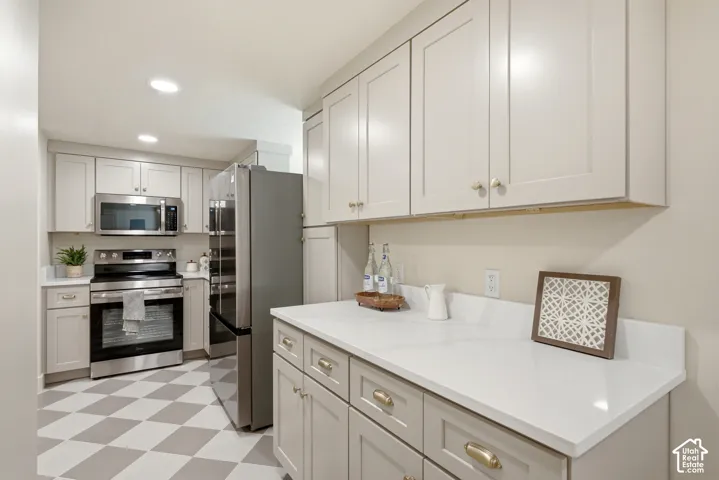 Recessed lighting illuminating light-toned cabinetry, stainless steel appliances, white countertops, and a distinct checkered floor pattern