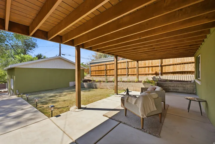 Covered concrete patio with exposed wood ceiling beams, adjacent to a grassy yard with perimeter fencing and tiered planters