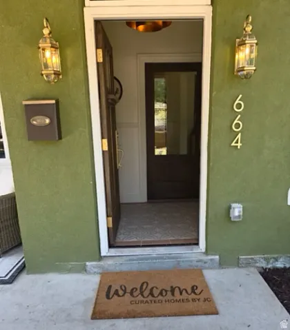 Green stucco exterior with dual brass-finish wall lanterns, a dark wood-finish entry door, and a patterned tile foyer