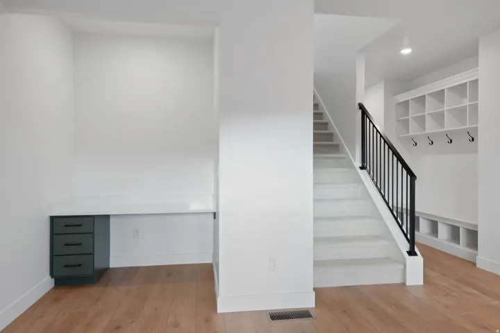 Built-in desk with solid surface countertop, wood-finish flooring, carpeted staircase with dark metal balusters, and built-in mudroom storage with hooks and cubbies