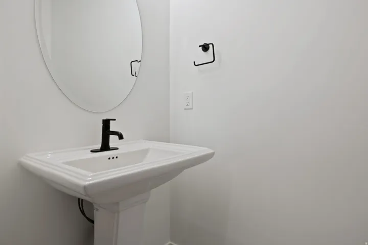 Pedestal sink with a matte black single-handle faucet, accompanied by an oval frameless mirror and a coordinating matte black towel ring
