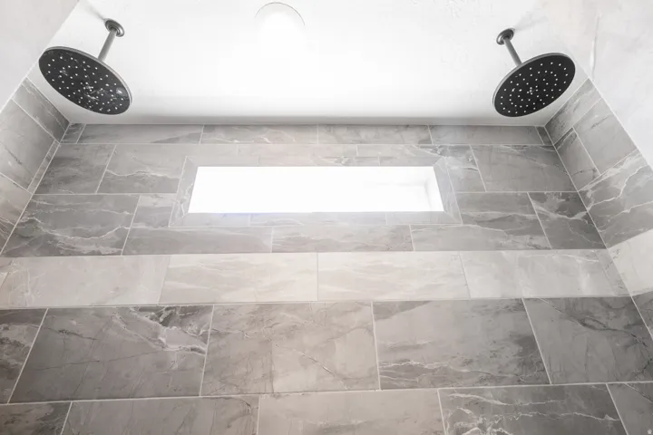 Dual ceiling-mounted showerheads with a matte black finish, textured gray tile walls, and a recessed wall niche