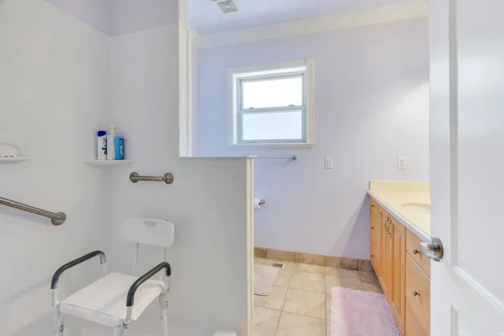 Primary suite bathroom with walk-in shower with grab bars, single vanity with light-tone countertop, and tiled flooring