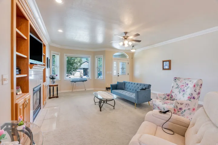 Built-in entertainment center with integrated shelving, fireplace with tile surround, bay window, crown molding, and ceiling fan with integrated lighting