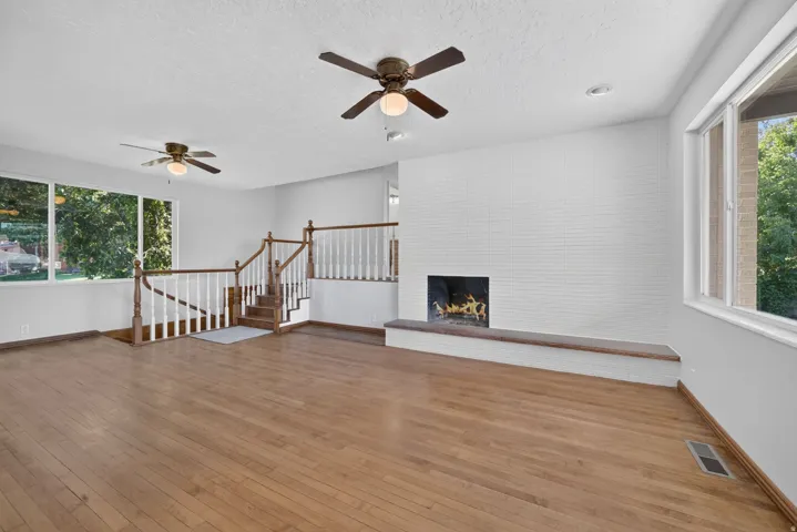 Wood-finish flooring, white brick fireplace, multiple large windows, wood staircase with white balusters, and dual ceiling fans