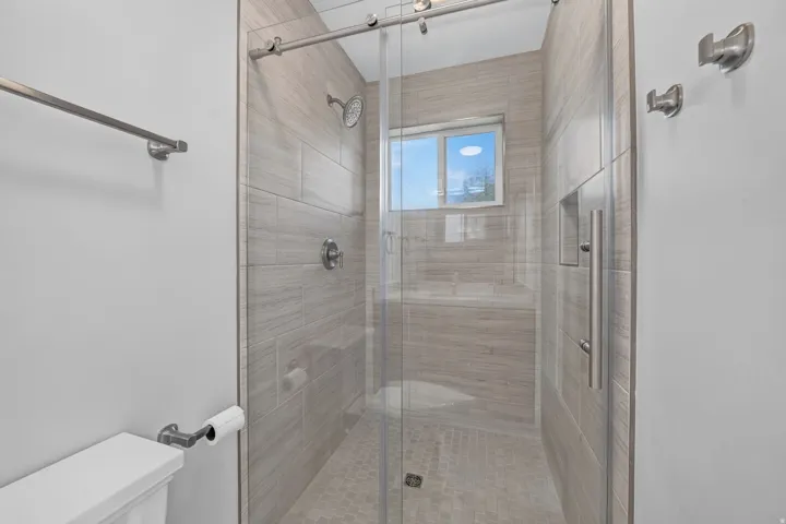 Glass-enclosed shower with full-height tile surround, built-in recessed shelf, and natural light from a privacy window