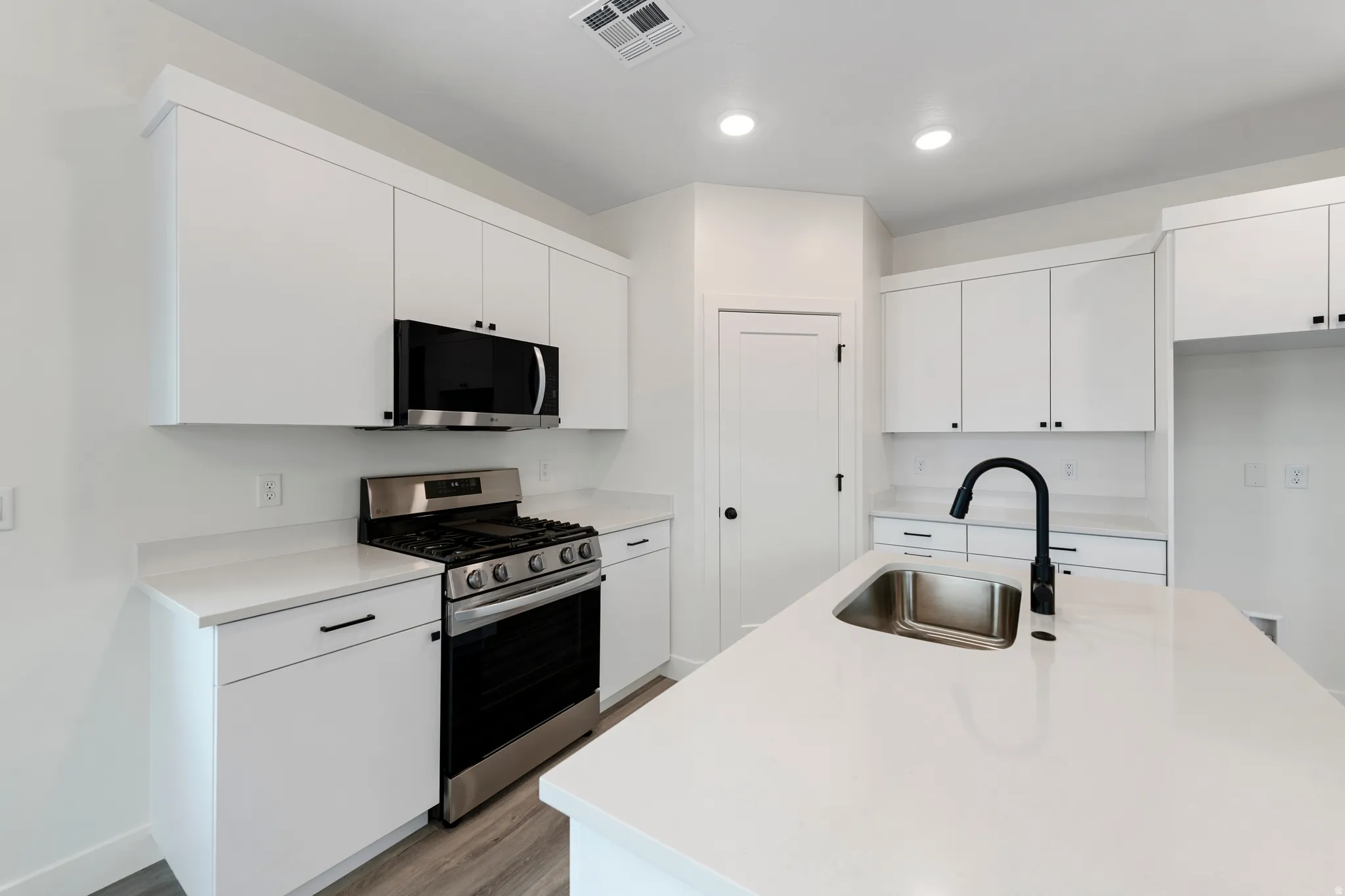 Bright kitchen featuring white cabinetry, stainless steel appliances, a center island with sink, black faucet, and wood-finish flooring