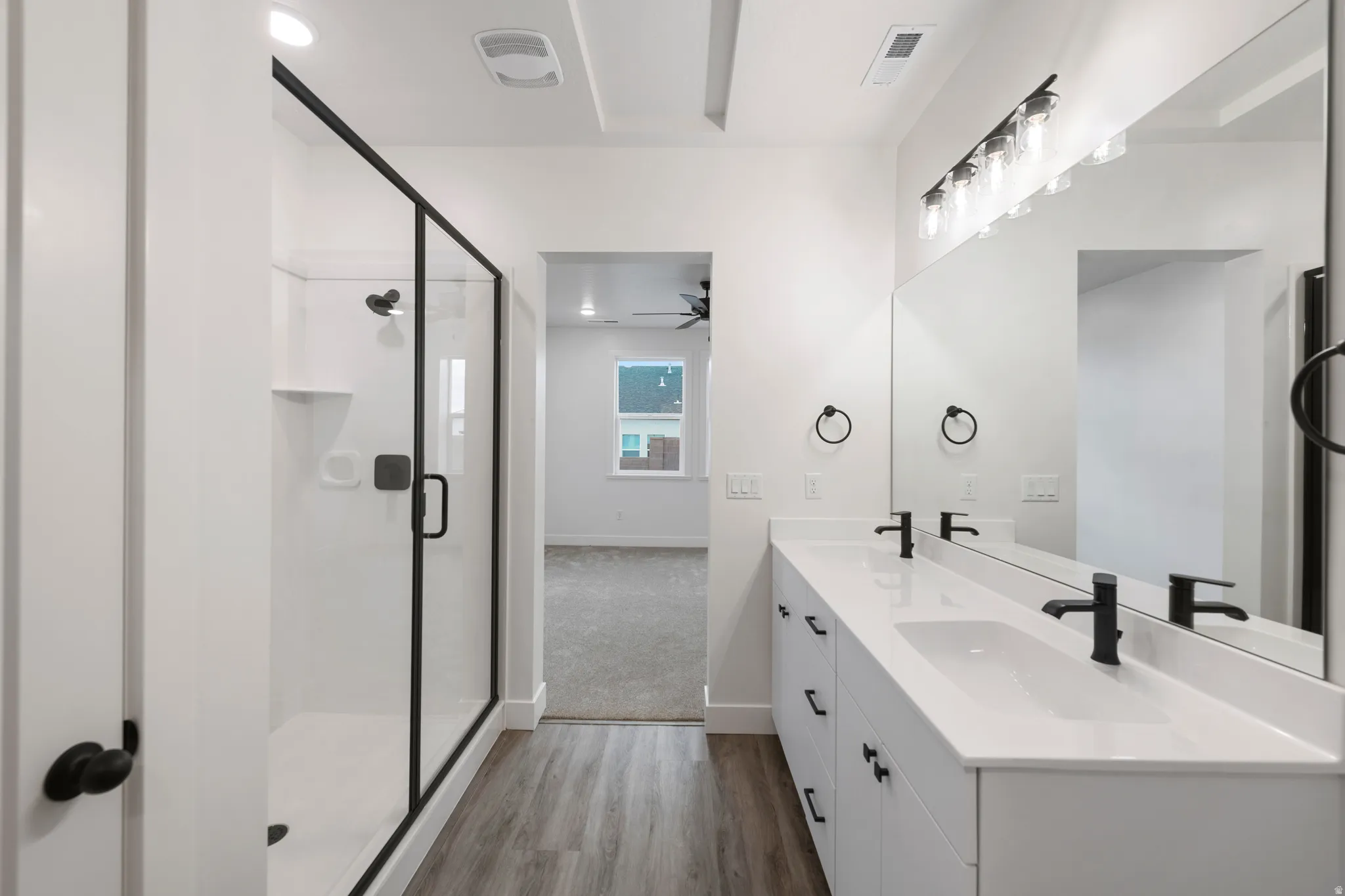 Dual vanity with undermount sinks and matte black fixtures, full-width mirror, wood-finish flooring, and a glass-enclosed shower with matte black framing