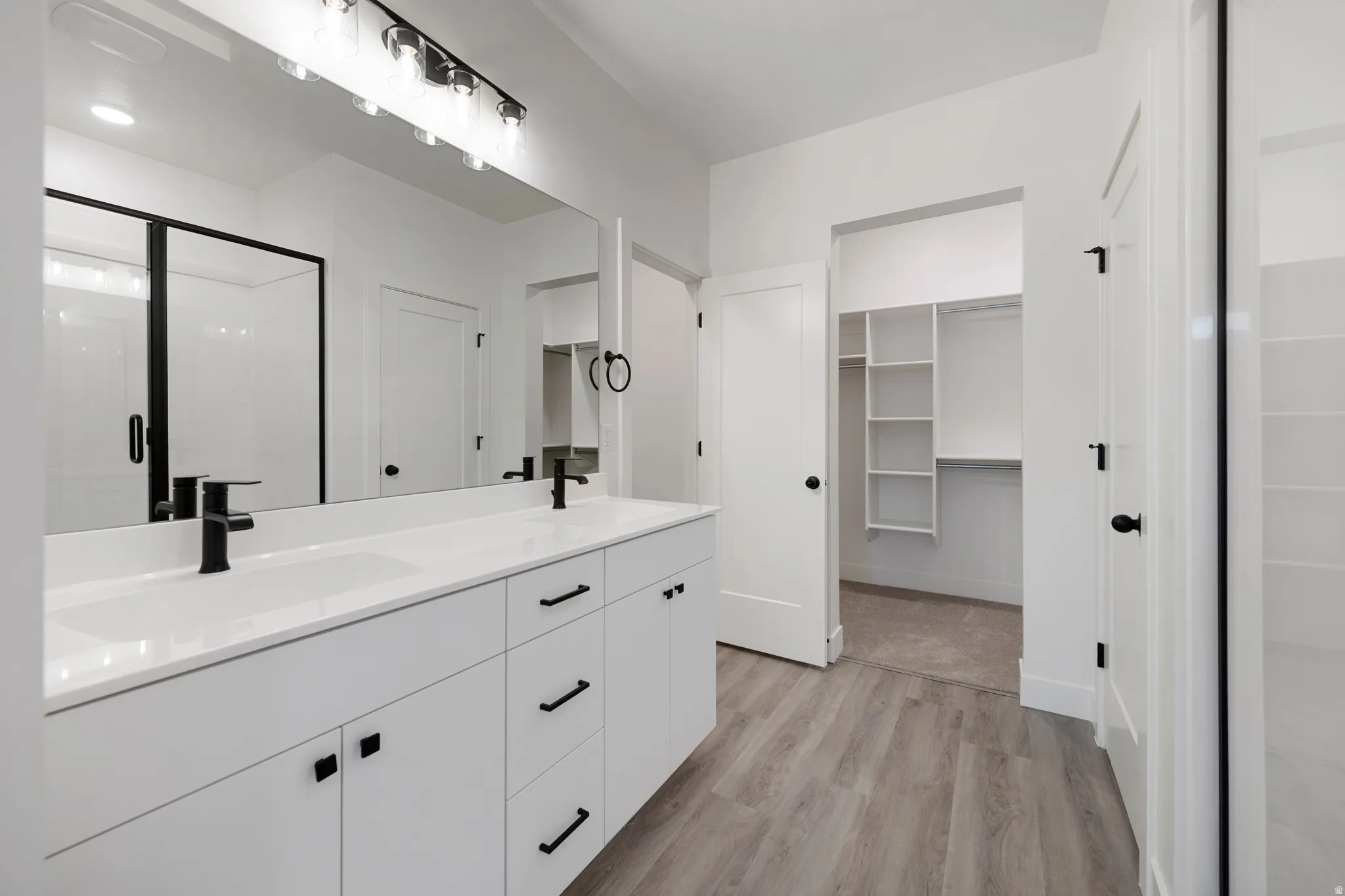 Dual vanity with white cabinetry and matte black hardware, modern frameless mirror, wood-finish flooring, and a walk-in closet with built-in shelving