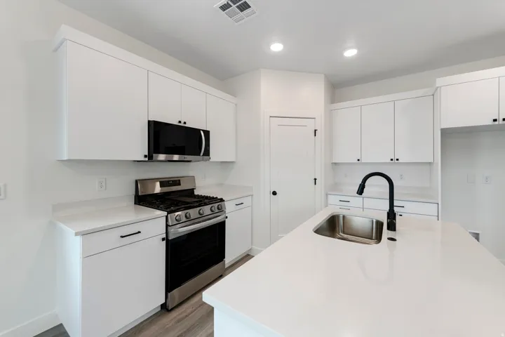 Bright kitchen featuring white cabinetry, stainless steel appliances, a center island with sink, black faucet, and wood-finish flooring