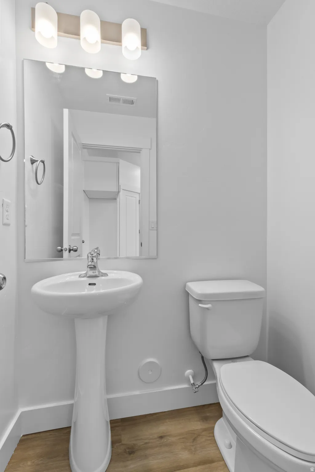 Pedestal sink with chrome faucet, wall-mounted mirror, brushed nickel light fixture with three sconces, toilet, and wood-finish flooring