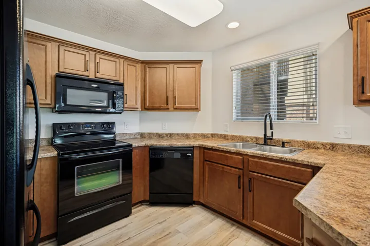 Kitchen featuring light wood-finish cabinetry, dark countertops, wood-finish flooring, stainless steel double basin sink with a matte black faucet, and black appliances
