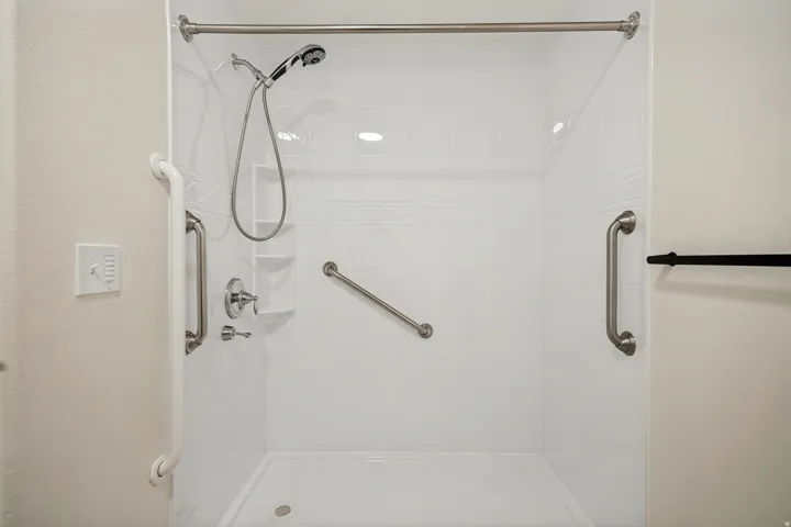 Walk-in shower with a handheld showerhead, multiple grab bars, recessed lighting, and built-in shelving