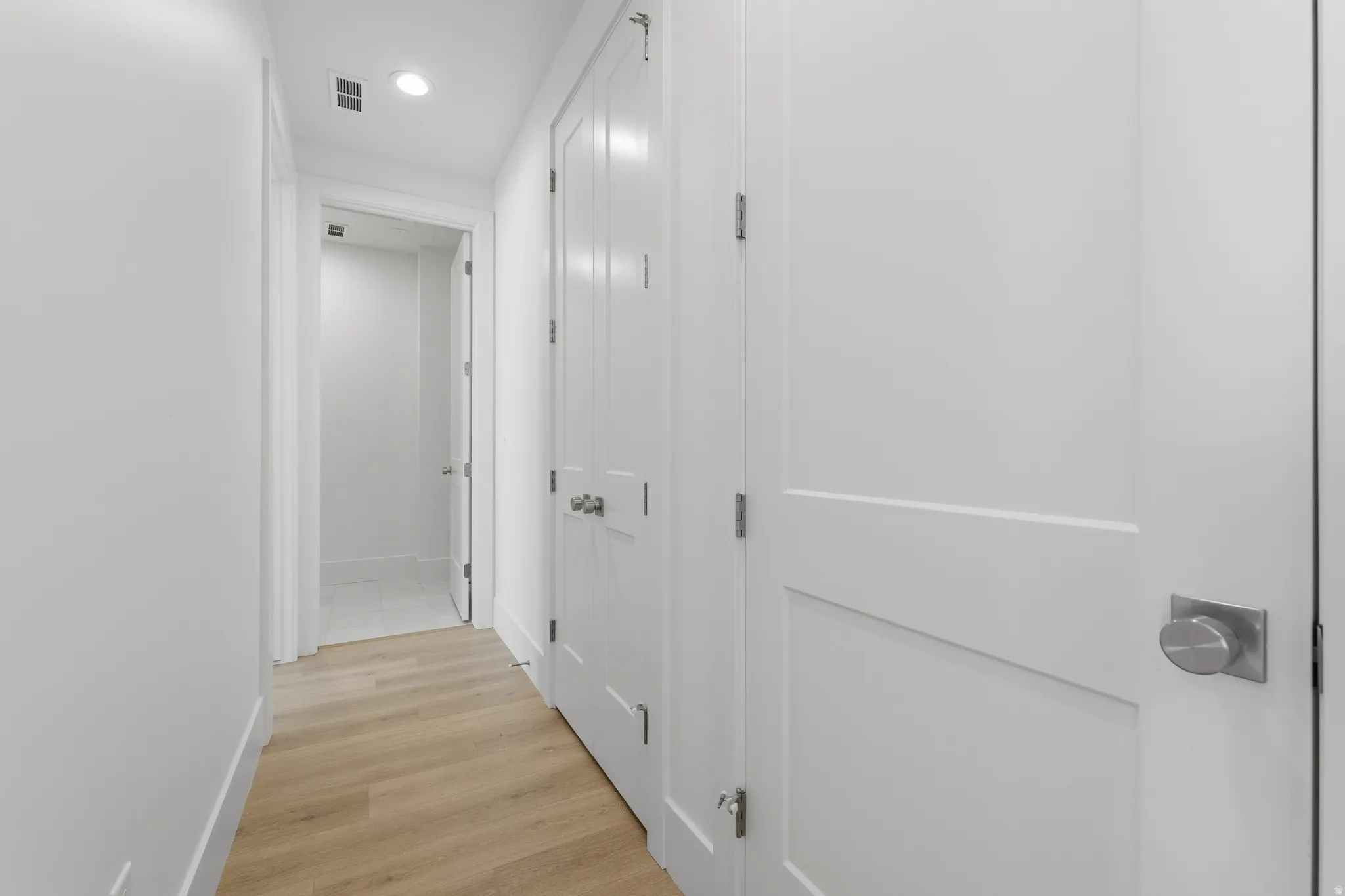 Hallway with wood-finish flooring, white walls, recessed lighting, and panel doors with brushed nickel hardware