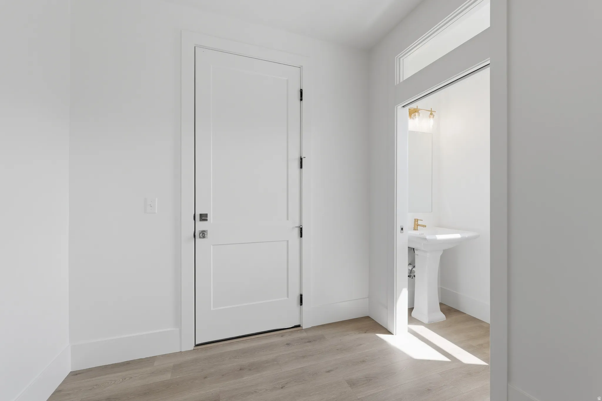 Wood-finish flooring throughout, white paneled door, and recessed powder room with a pedestal sink, gold-tone faucet, and vanity lighting