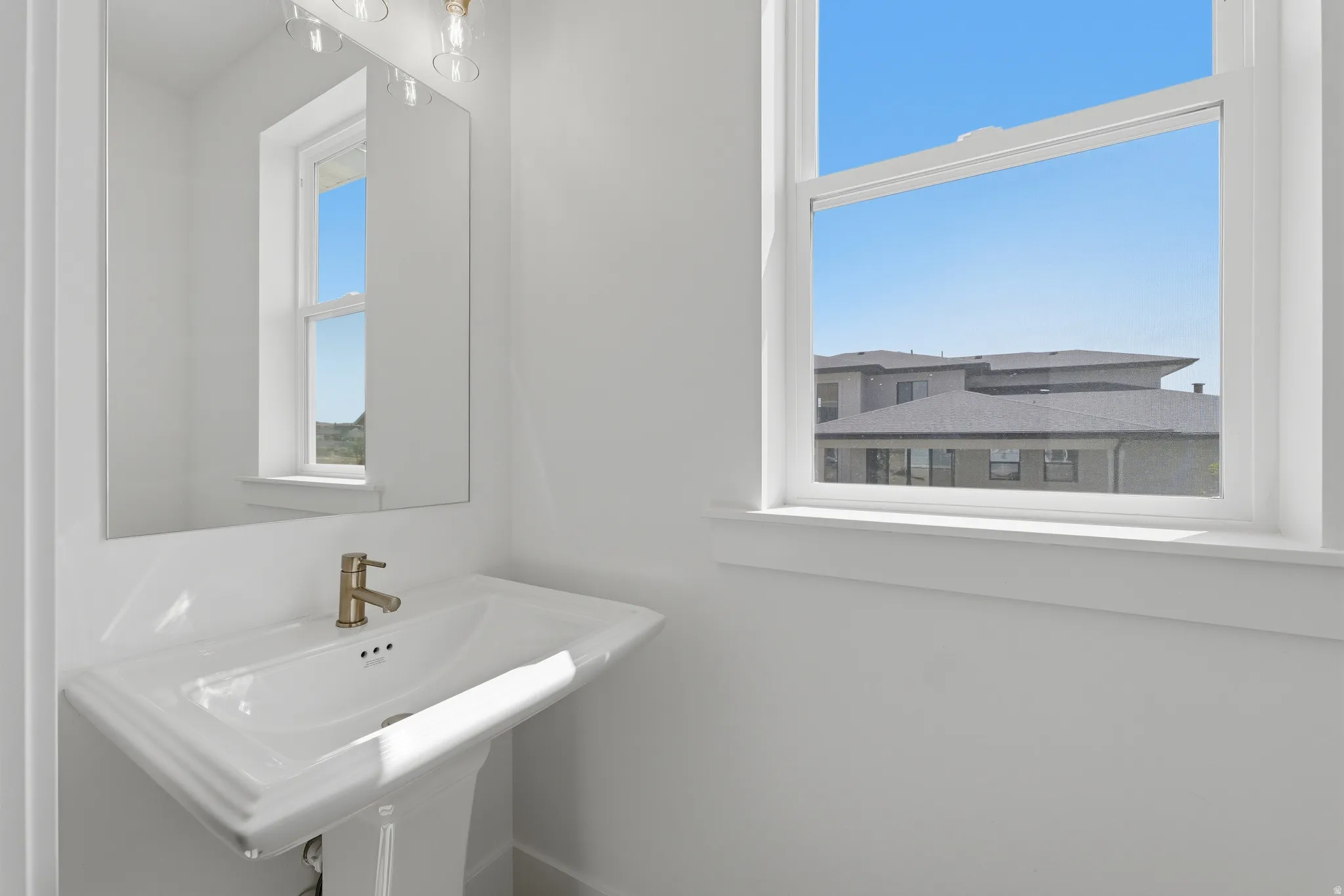 Pedestal sink with brass-finish faucet, rectangular wall mirror, and window with a white frame