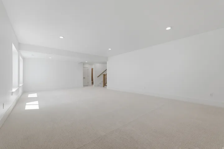 Spacious room featuring neutral carpeting, recessed lighting, multiple windows, and a staircase with wood railings
