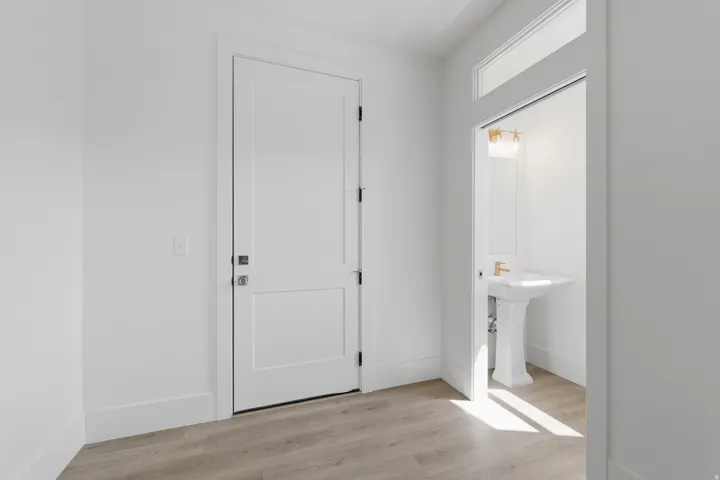 Wood-finish flooring throughout, white paneled door, and recessed powder room with a pedestal sink, gold-tone faucet, and vanity lighting