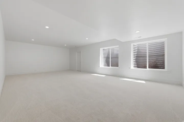 Spacious room featuring light-toned carpet, recessed lighting, two windows with window wells, and a white interior door
