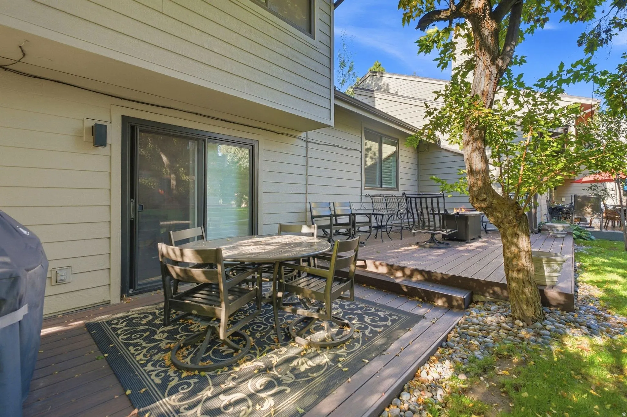 Multi-level wood-finish deck with exterior siding, sliding glass door, mature trees, and landscaped rock beds