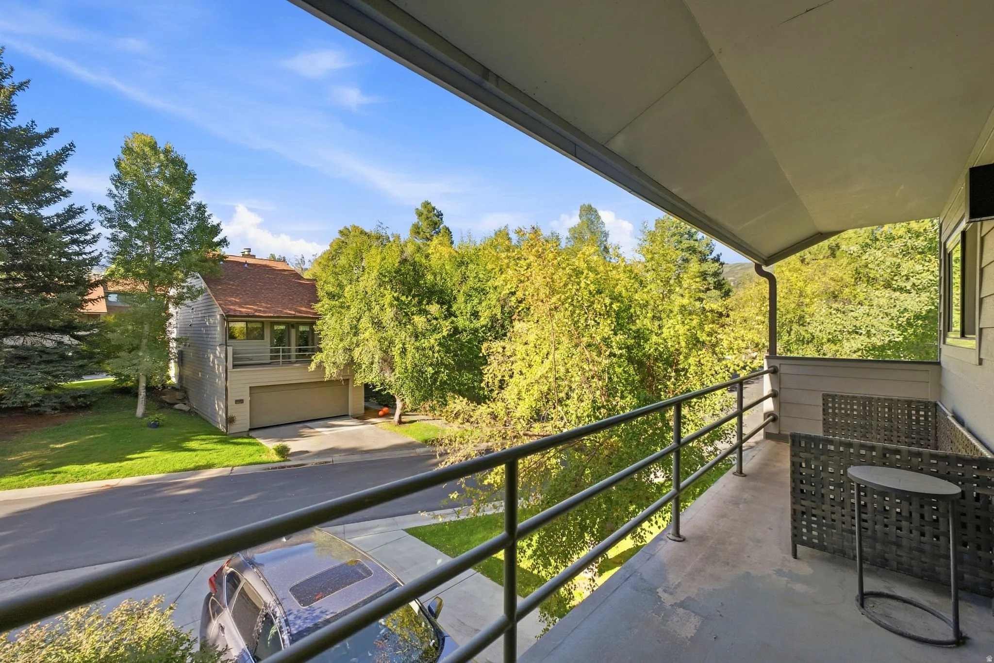 Covered exterior balcony with metal railing, an adjacent residential structure, mature landscaping, and an asphalt paved road