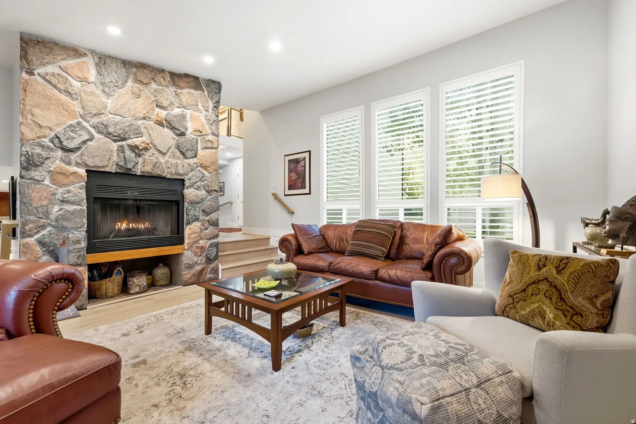 Stone-clad fireplace with a raised hearth, wood-finish flooring, and multiple vertical windows featuring plantation shutters