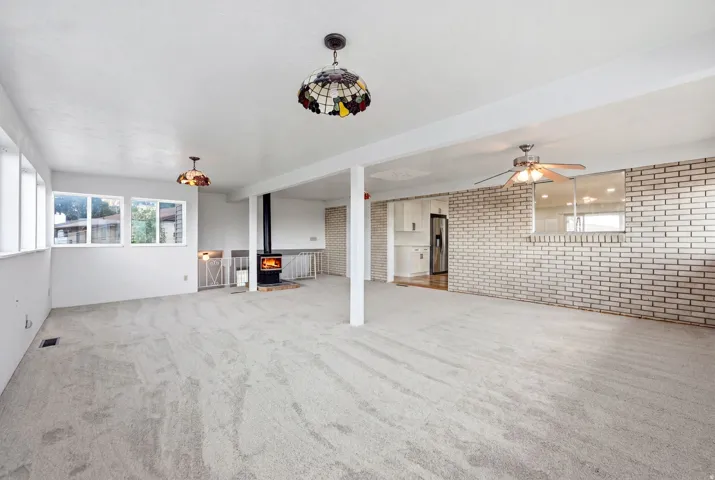 Spacious room featuring light-toned carpeting, multiple windows, a standalone wood-burning stove, and a ceiling fan