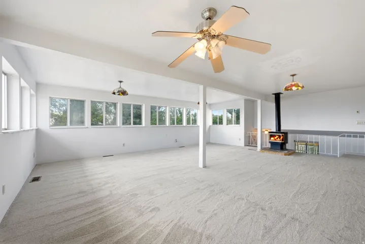 Spacious room featuring light-toned carpeting, multiple windows, a ceiling fan with integrated lighting, and a freestanding wood-burning stove with a black pipe