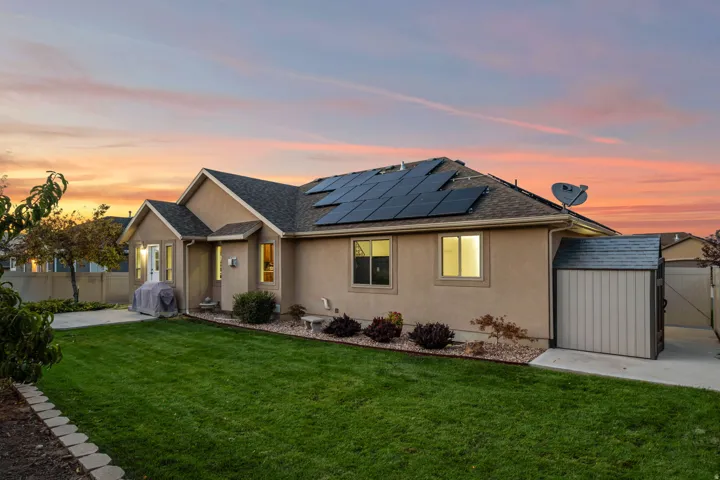 Expansive green lawn, stucco exterior, multiple solar panels, private concrete patio, and a dedicated storage shed