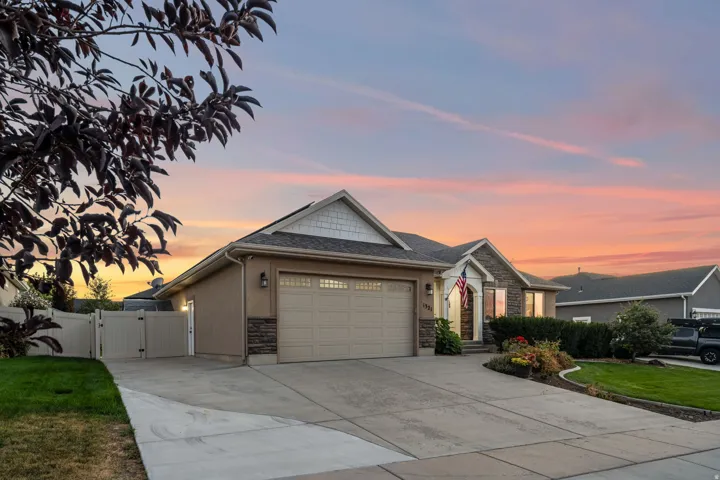 Stucco and stone facade with a two-car garage, mature landscaping, concrete driveway, and a white privacy fence