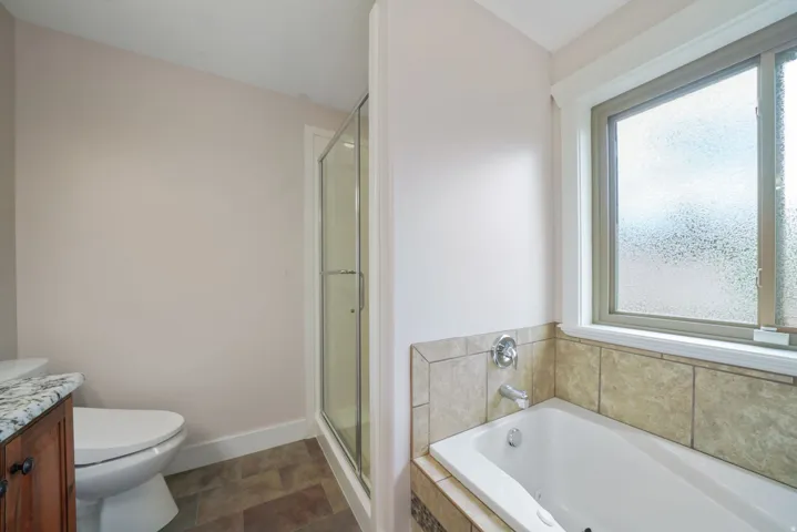 Soaking tub with tile surround, separate glass-enclosed shower, residential window with frosted glass, wood-finish vanity with stone countertop, and tile flooring