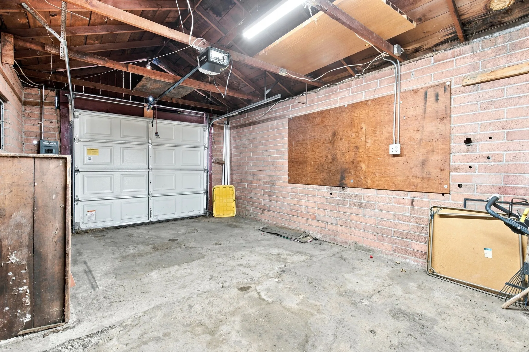 Single garage door with an automatic opener, exposed timber ceiling beams, brick walls, and a concrete floor
