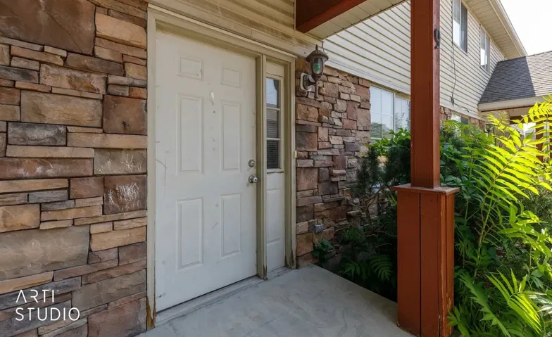 Stacked stone exterior with a paneled entry door, concrete porch, wood support column, and exterior lighting fixture