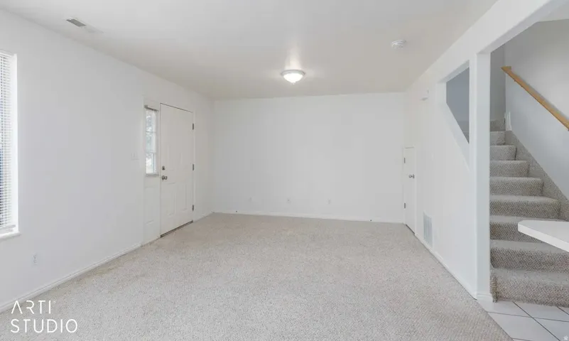 Carpeted living space featuring a window with blinds, a white door, and a staircase with a wood-finish handrail