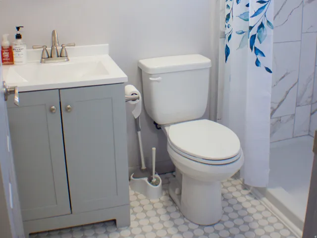 Window-lit bathroom featuring a light gray vanity with an integrated sink, round wall mirror, white penny tile flooring, and tiled shower.
