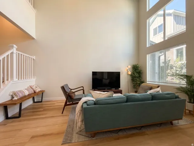 Expansive living area with high ceilings, large windows with blinds, light wood-finish flooring, and white painted stair railing