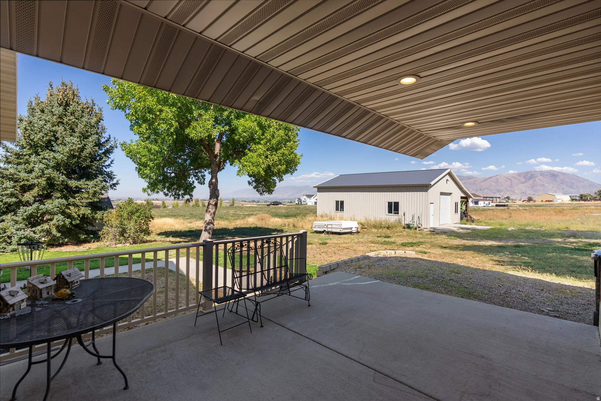 Covered patio with integrated lighting and railing, concrete patio surface, and a detached auxiliary building with a roll-up door