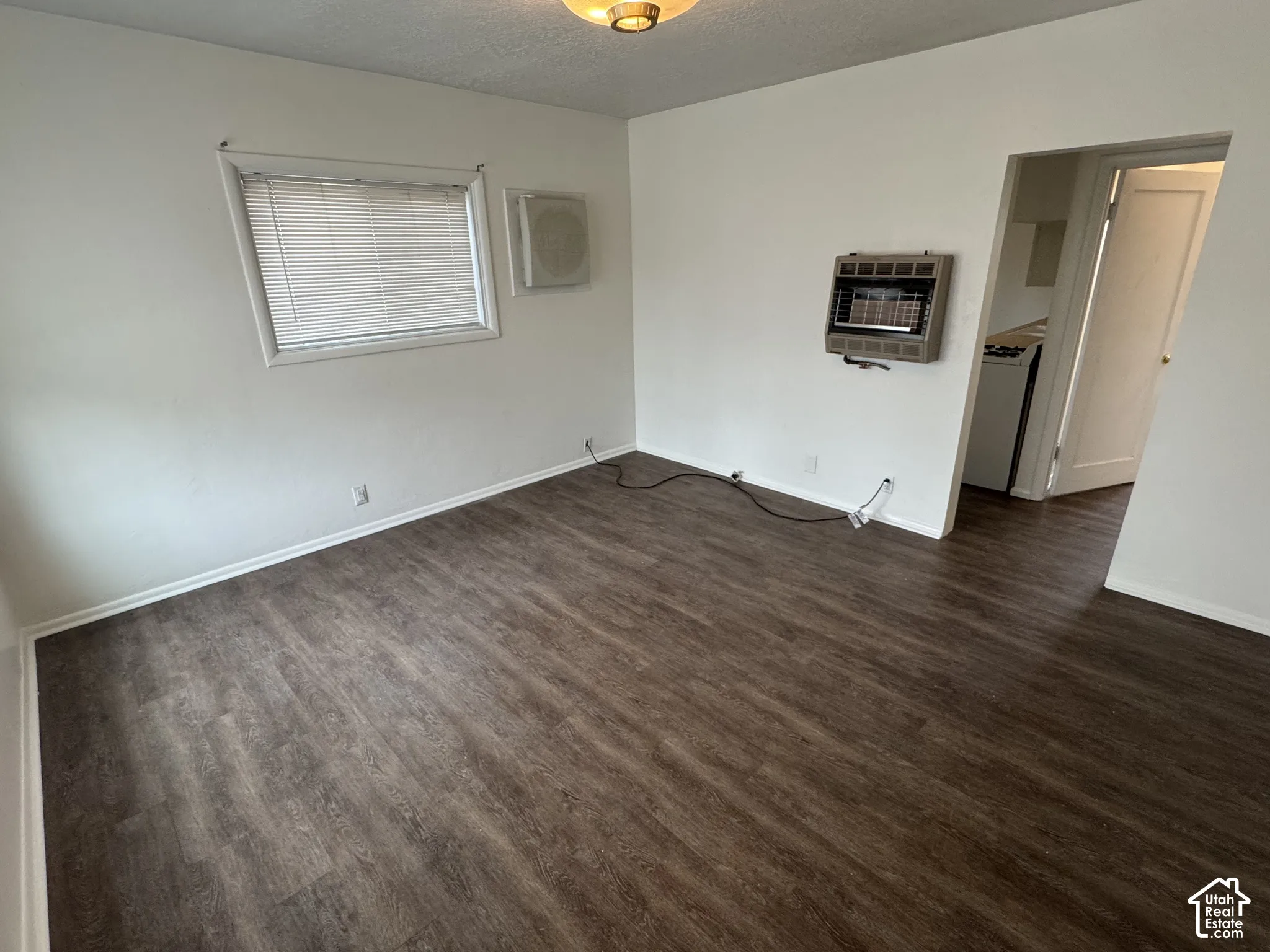 Wood-finish flooring throughout, white painted walls, window with blinds, wall-mounted heater, and ceiling-mounted light fixture