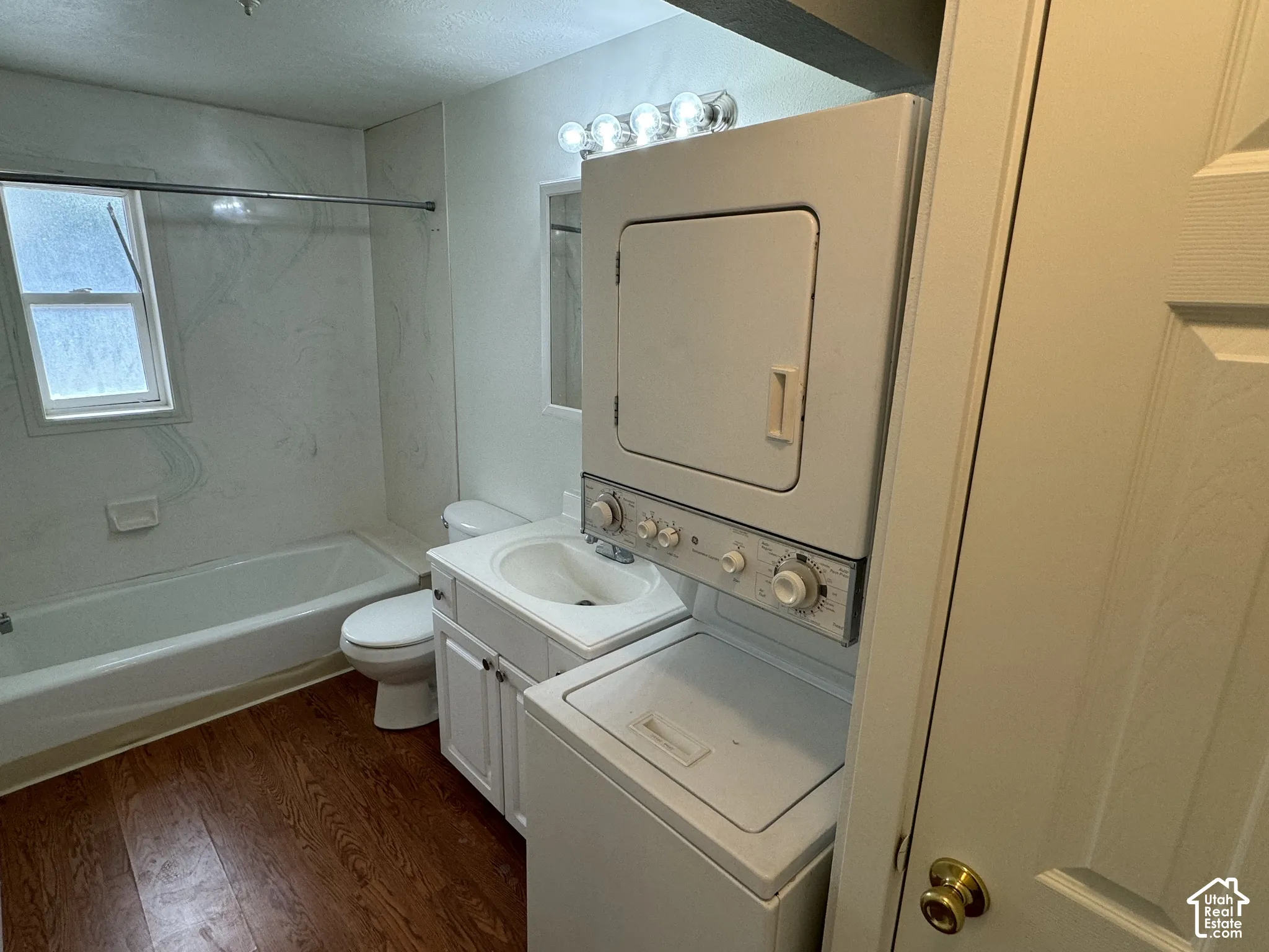 Wood-finish flooring, white vanity with integrated sink, toilet, tub with shower kit, and a stacked washer and dryer